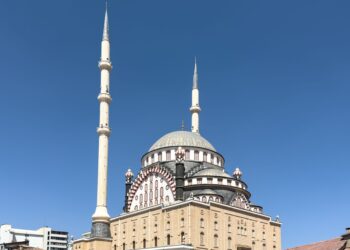 Açık mavi gökyüzü altında iki minareli ve büyük kubbeleri olan görkemli bir cami.