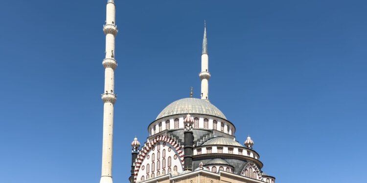 Açık mavi gökyüzü altında iki minareli ve büyük kubbeleri olan görkemli bir cami.