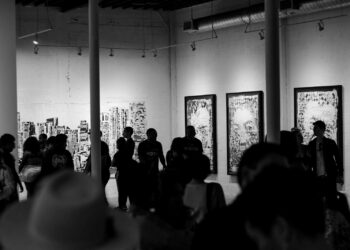 Siyah beyaz fotoğrafta sanat galerisindeki tablo ve eserleri inceleyen kalabalık.