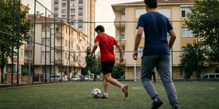 Sokak aralarındaki halı sahasında futbol oynayan gençlerin neşeli mücadelesi.