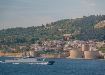 Denizde ilerleyen bir savaş gemisi ve kıyıda tarihi kale yapısı