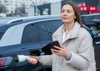 Şarj istasyonunda elektrikli otomobilini şarj ederken telefonuna bakan bir kadın